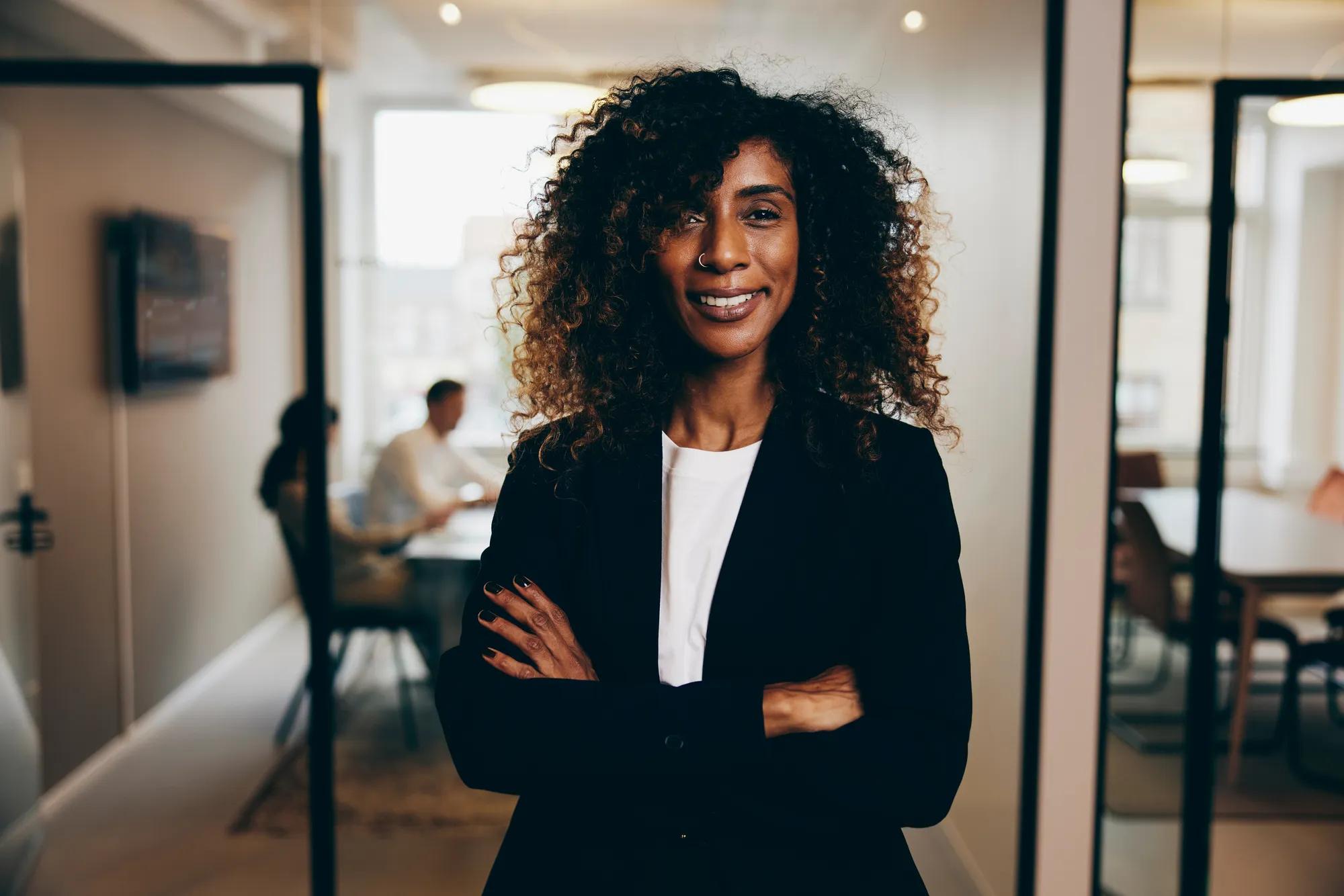 Smiling businesswoman standing with her arms crossed in an office with coworkers having a meeting in the background