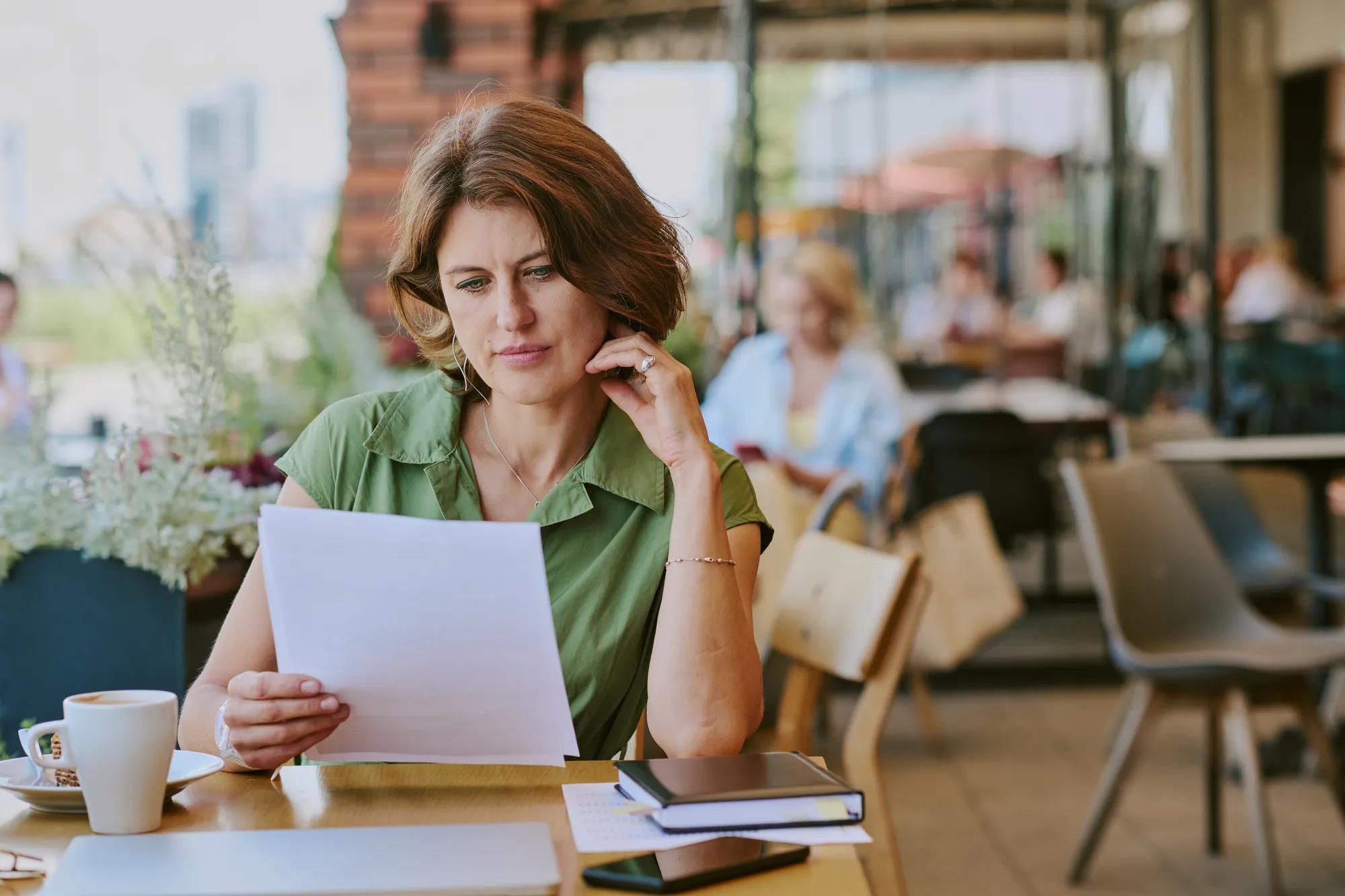 Caucasian middle aged woman reading documents while sitting at outdoor cafe table, holding paper in one hand, thoughtful expression, coffee cup and notebooks on table