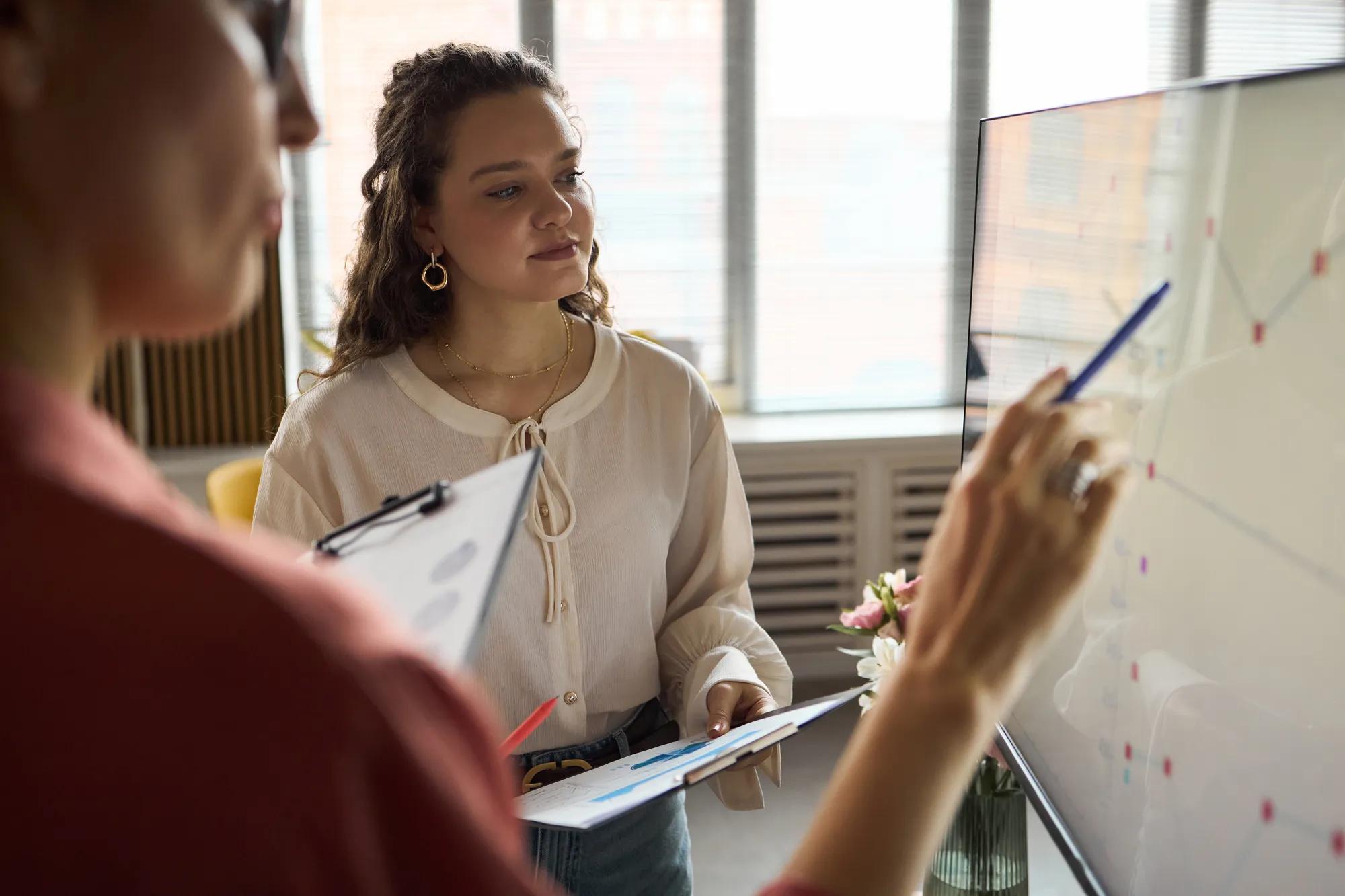 Young adult Caucasian woman listening attentively while colleague explaining data on whiteboard during business meeting in modern office, both holding documents and discussing strategy