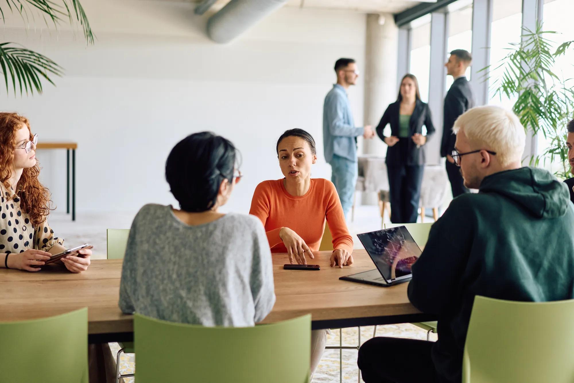 A diverse team of business professionals engaged in a discussion around a conference table in an office, while their colleagues collaborate in the background. 